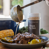 A jar of Clean Monday Meals Clean Au Jus seasoning stands on a table beside a plated cooked meat dish, corn on the cob, and a ladle pouring sauce into the plate in a real kitchen setting.