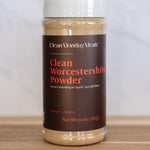 A 10 oz plastic jar of Clean Monday Meals Clean Worcestershire Powder on a wooden surface with a light marble background.