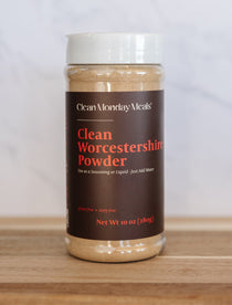 A 10 oz plastic jar of Clean Monday Meals Clean Worcestershire Powder on a wooden surface with a light marble background.