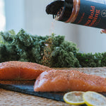 A bottle of Bold Seasoning is being tilted to sprinkle seasoning onto two raw salmon fillets on a dark slate board, with kale and lemon slices in the background.