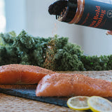 A bottle of Bold Seasoning is being tilted to sprinkle seasoning onto two raw salmon fillets on a dark slate board, with kale and lemon slices in the background.