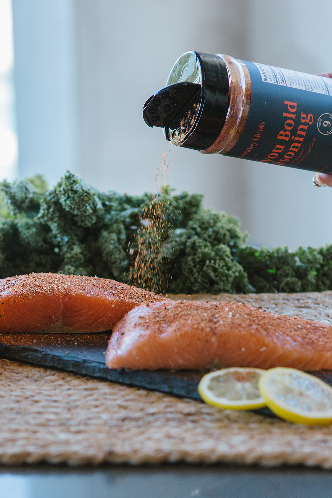 A bottle of Bold Seasoning is being tilted to sprinkle seasoning onto two raw salmon fillets on a dark slate board, with kale and lemon slices in the background.