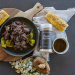 A styled food scene on a dark gray surface showing a bowl of shredded pulled meat with green peppers, a corn cob, a small cup of brown sauce, a plate with mashed potato and chopped onions, and a jar labeled Clean All Jus standing in the center.