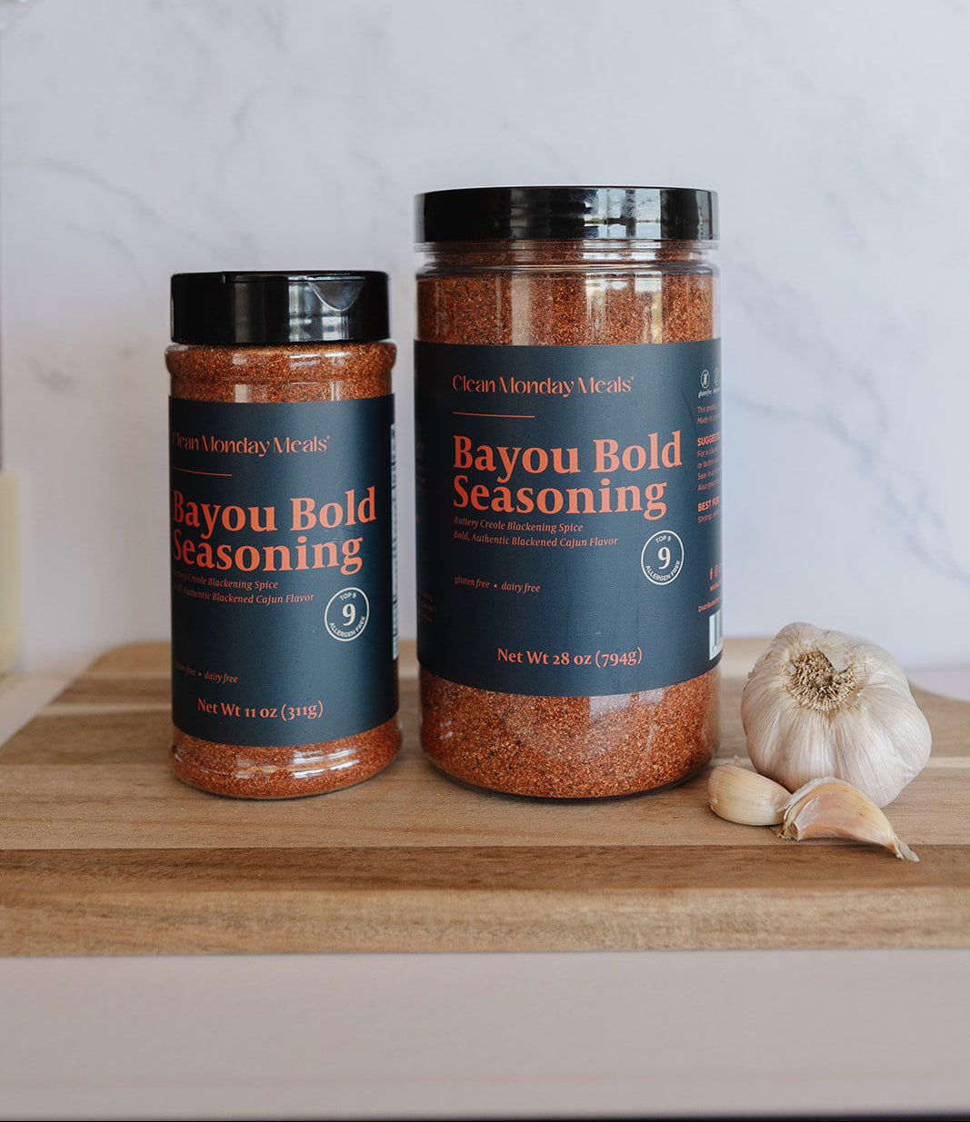 Two jars of Claymonkey Smoke Bayou Bold Seasoning displayed on a wooden cutting board next to a whole garlic bulb, with a white marble backsplash in the background.