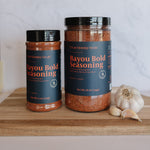 Two jars of Claymonkey Smoke Bayou Bold Seasoning displayed on a wooden cutting board next to a whole garlic bulb, with a white marble backsplash in the background.
