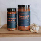 Two jars of Claymonkey Smoke Bayou Bold Seasoning displayed on a wooden cutting board next to a whole garlic bulb, with a white marble backsplash in the background.