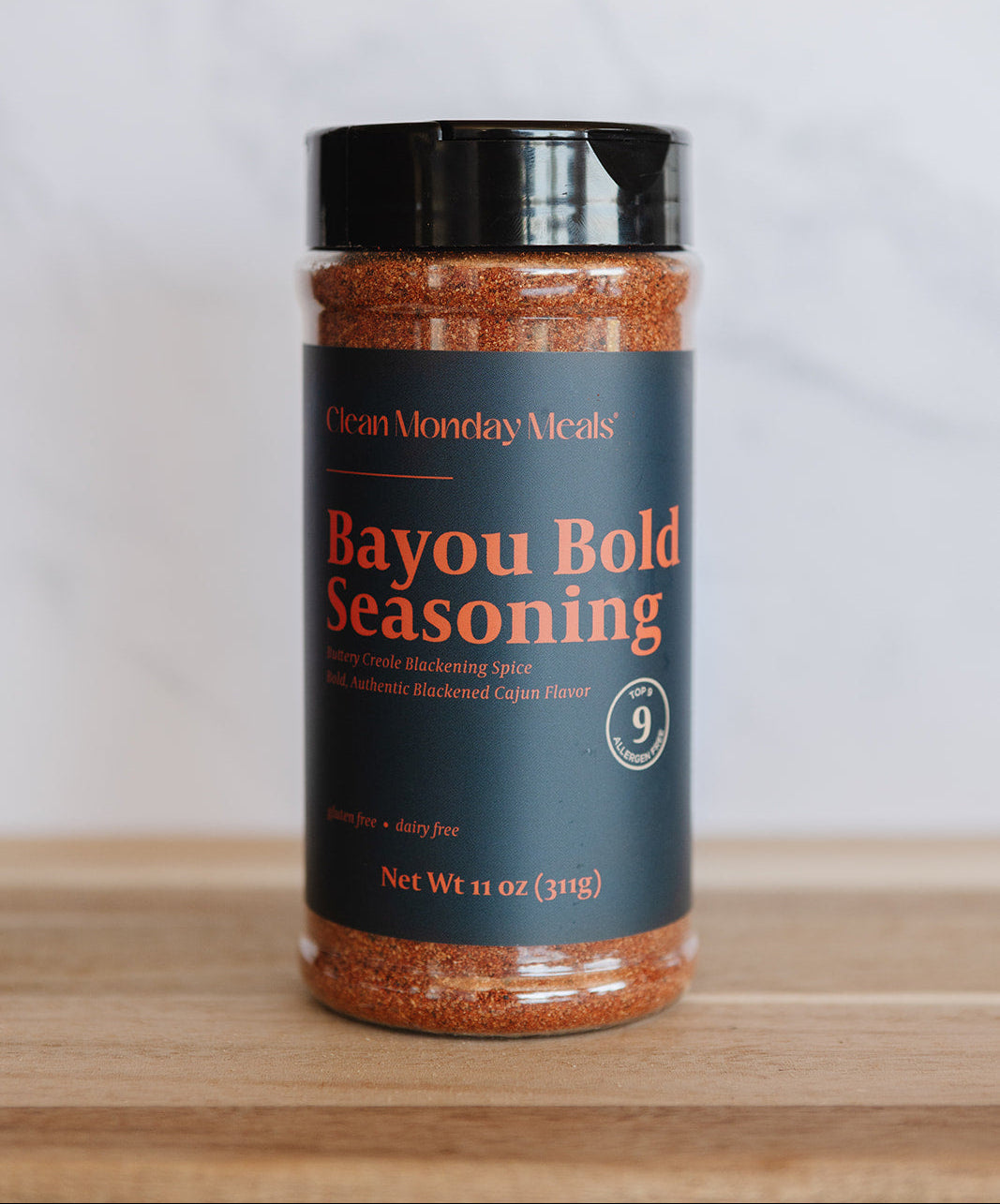 A bottle of Bayou Bold Seasoning by Clean Monday Meals, featuring a dark teal label with orange text and a black lid, set on a wooden surface against a light gray background.