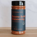 A bottle of Bayou Bold Seasoning by Clean Monday Meals, featuring a dark teal label with orange text and a black lid, set on a wooden surface against a light gray background.