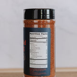 A clear plastic shaker bottle filled with reddish-brown seasoning, featuring a black lid and a dark label showing nutrition facts, set on a wooden surface against a light gray background.