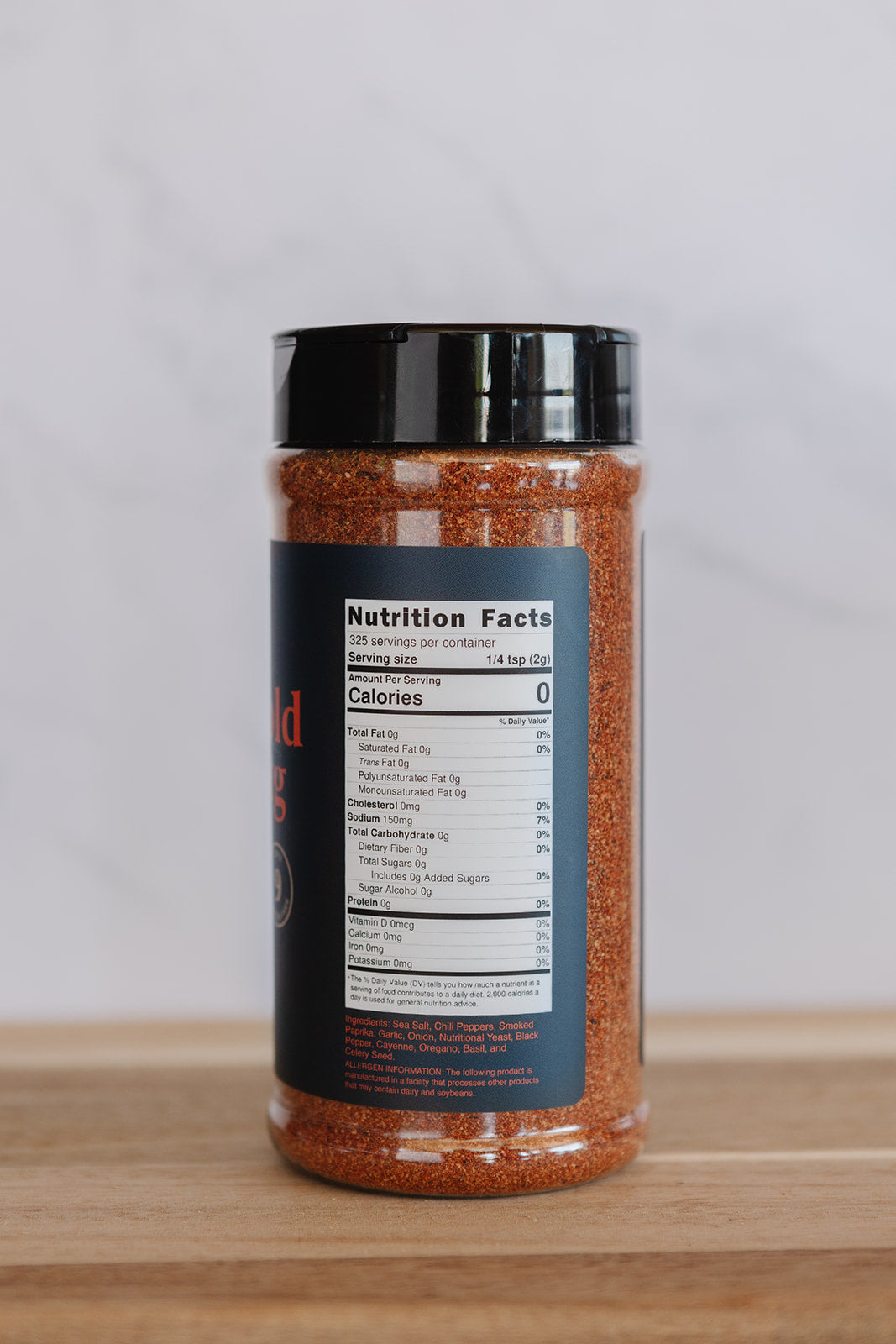 A clear plastic shaker bottle filled with reddish-brown seasoning, featuring a black lid and a dark label showing nutrition facts, set on a wooden surface against a light gray background.
