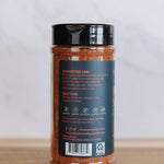 A clear plastic jar of seasoning with a black lid, showing the back label with text and icons, placed on a wooden surface against a light gray marble background.