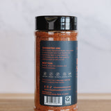 A clear plastic jar of seasoning with a black lid, showing the back label with text and icons, placed on a wooden surface against a light gray marble background.