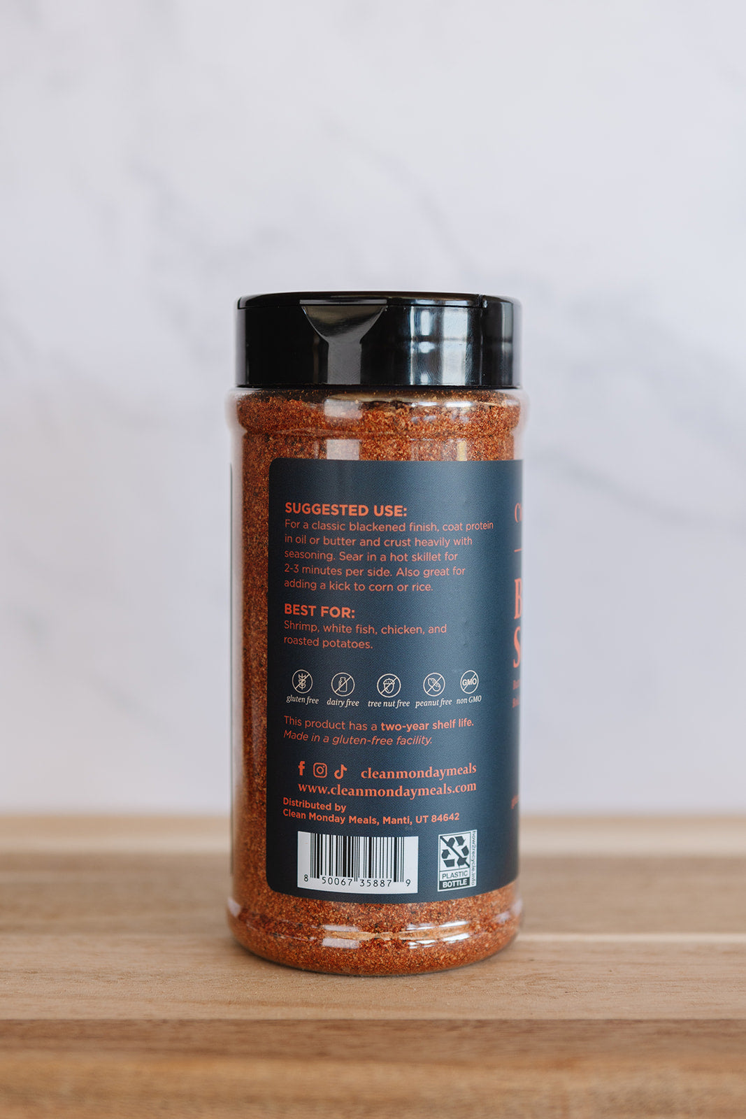 A clear plastic jar of seasoning with a black lid, showing the back label with text and icons, placed on a wooden surface against a light gray marble background.