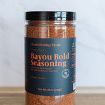 A jar of Ocean Monday Meals Bayou Bold Seasoning on a wooden surface with a light gray marble background. The product is in a clear plastic jar showing the reddish-brown seasoning inside, with a black lid and a dark navy label featuring orange and white text.