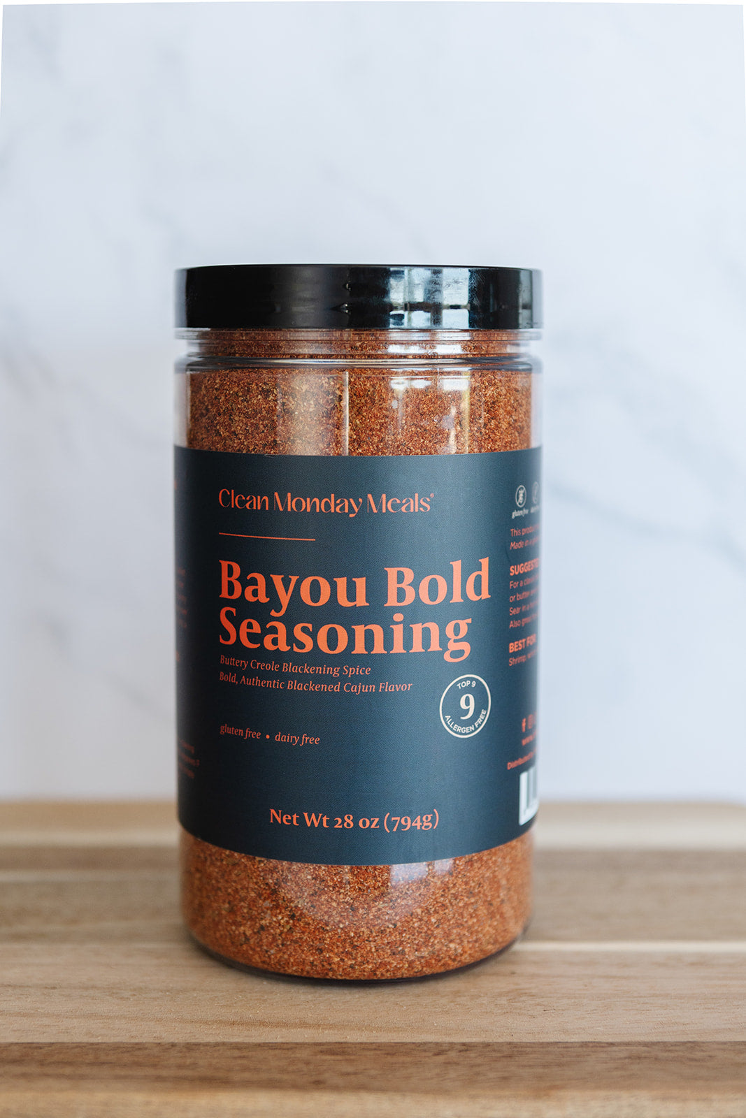 A jar of Ocean Monday Meals Bayou Bold Seasoning on a wooden surface with a light gray marble background. The product is in a clear plastic jar showing the reddish-brown seasoning inside, with a black lid and a dark navy label featuring orange and white text.