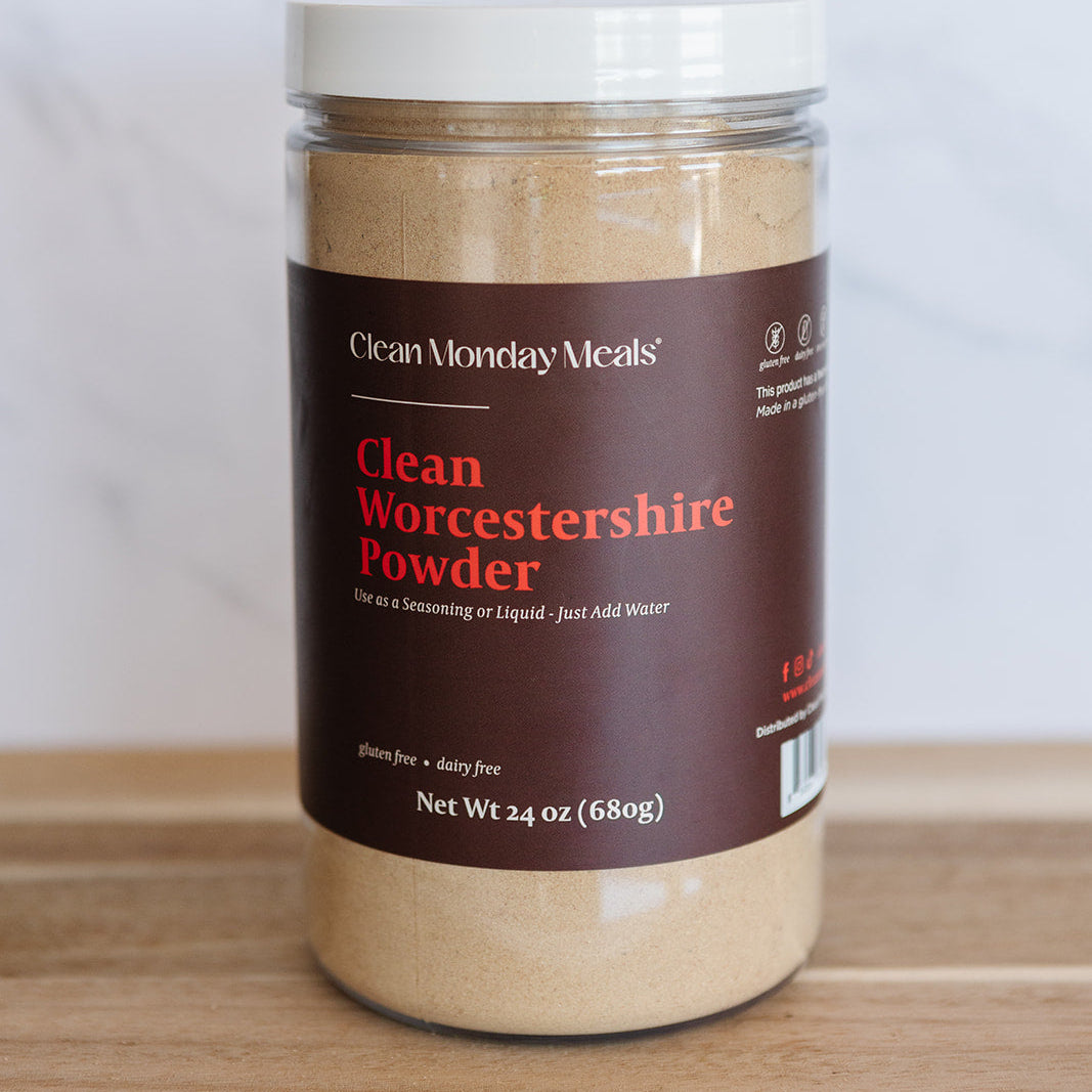 A jar of Clean Monday Meals Clean Worcestershire Powder on a wooden surface against a light marble background.