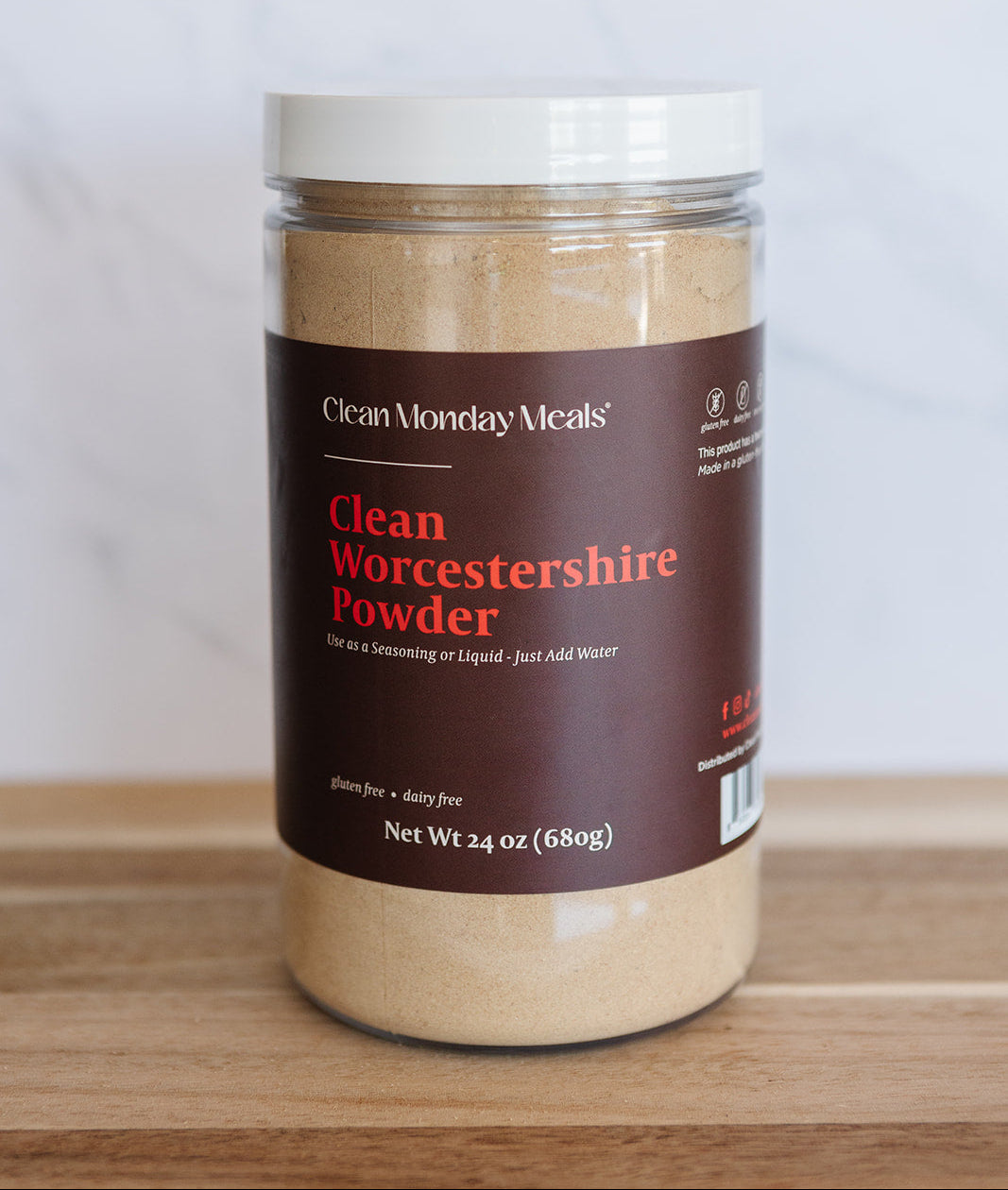 A jar of Clean Monday Meals Clean Worcestershire Powder on a wooden surface against a light marble background.