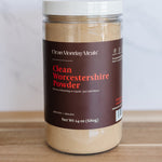 A jar of Clean Monday Meals Clean Worcestershire Powder on a wooden surface against a light marble background.