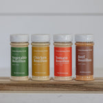 Four bottles of bouillon in different flavors on a wooden shelf with a white background