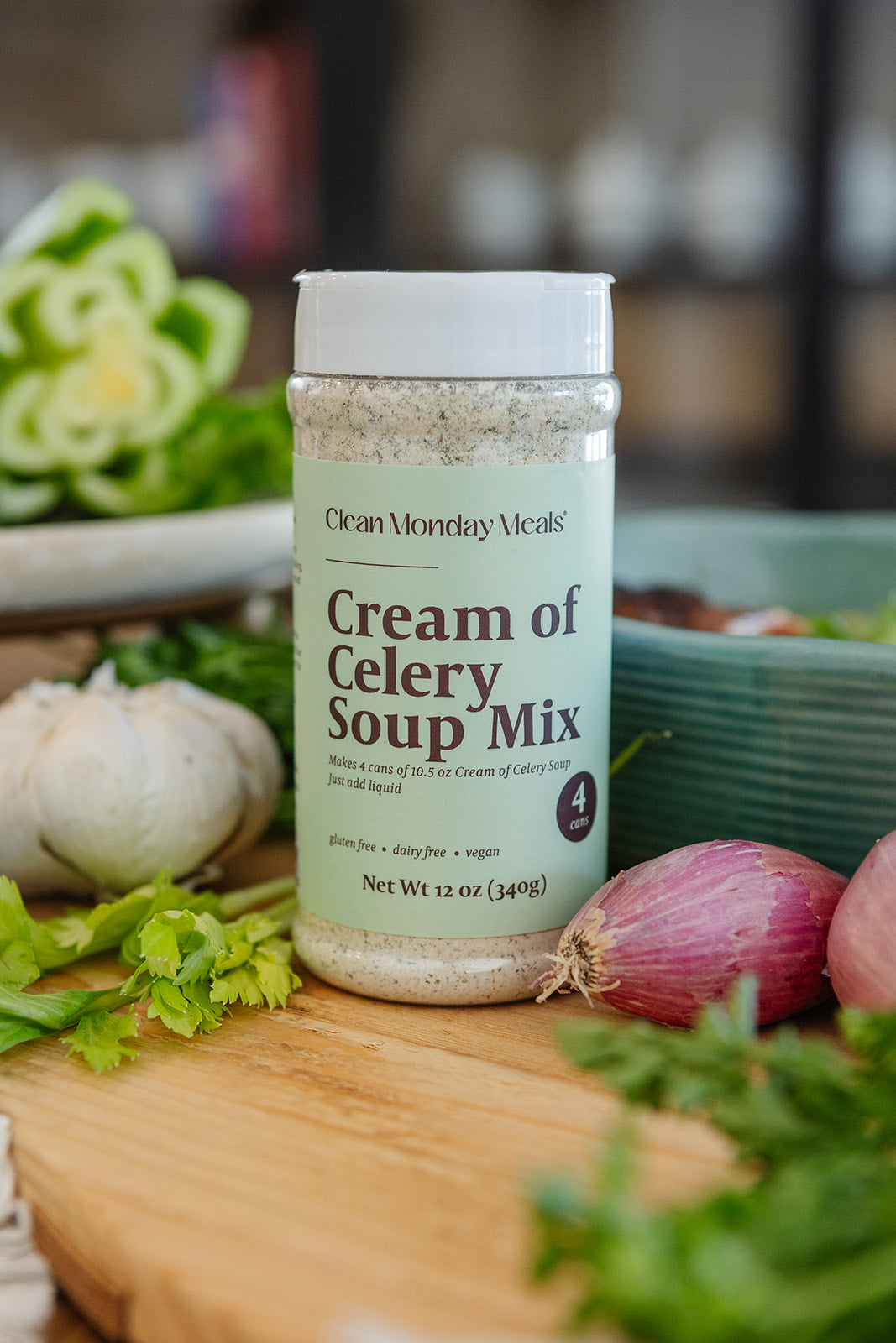 A bottle of Cream of Celery Soup Mix by Clean Monday Meals stands on a wooden cutting board surrounded by fresh ingredients like celery, garlic, red onions, and green beans in a kitchen setting.