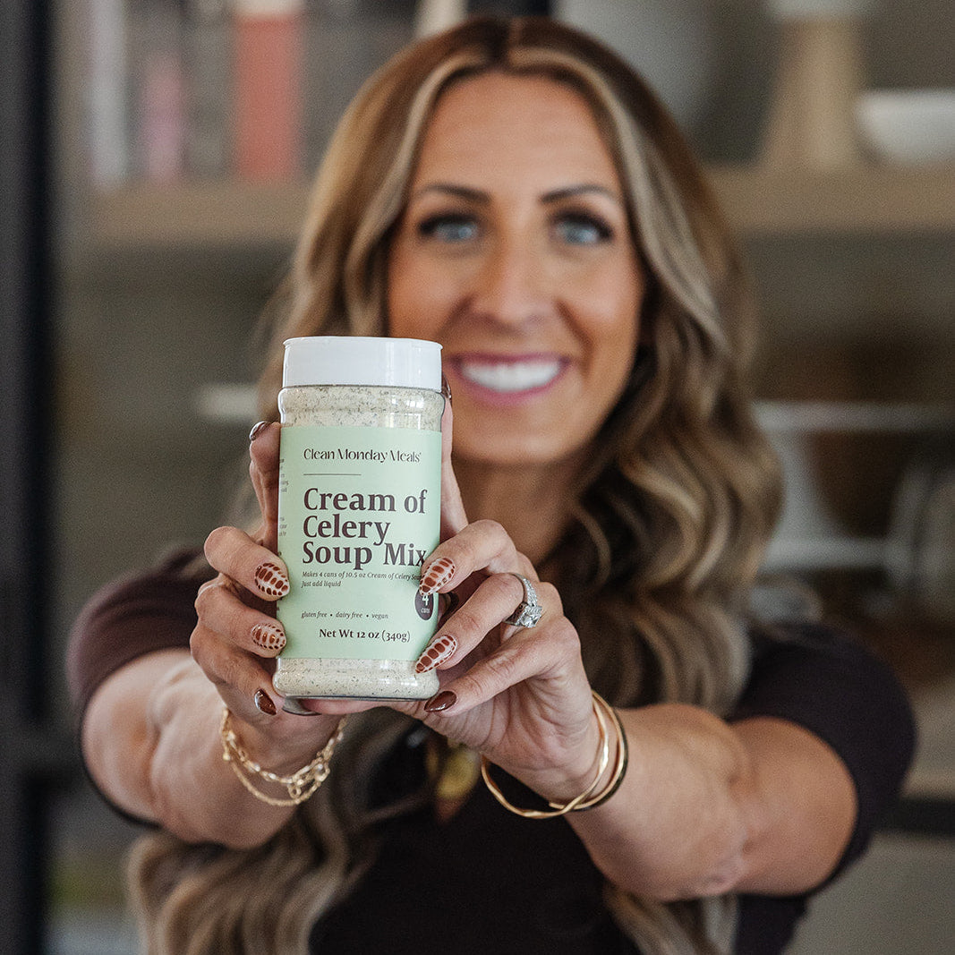 A smiling woman holds up a jar of Clean Monday Meals Cream of Celery Soup Mix towards the camera in a kitchen setting.