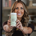 A smiling woman holds up a jar of Clean Monday Meals Cream of Celery Soup Mix towards the camera in a kitchen setting.