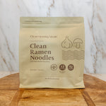 A stand-up pouch of Clean Ramen Noodles by Clean Monday Meals sits on a wooden board against a white marble-patterned background.