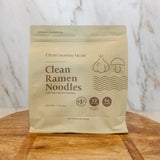 A stand-up pouch of Clean Ramen Noodles by Clean Monday Meals sits on a wooden board against a white marble-patterned background.