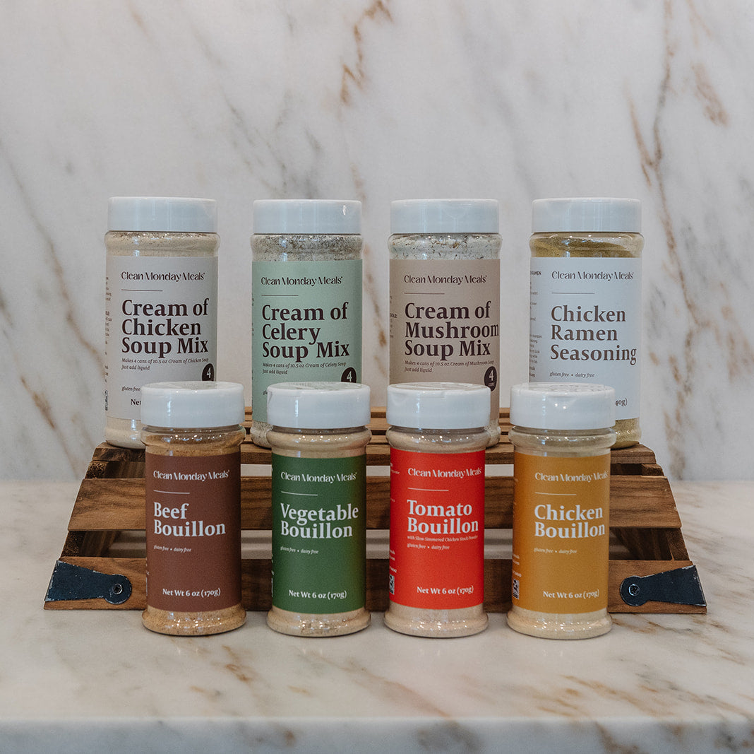 A set of eight seasoning and soup mix spice bottles from Clean Monday Meals, arranged on a wooden stand on a marble countertop with a marble backsplash. The bottles include Cream of Chicken Soup Mix, Cream of Celery Soup Mix, Cream of Mushroom Soup Mix, Chicken Ramen Seasoning, Beef Bouillon, Vegetable Bouillon, Tomato Bouillon, and Chicken Bouillon.