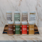 A set of eight seasoning and soup mix spice bottles from Clean Monday Meals, arranged on a wooden stand on a marble countertop with a marble backsplash. The bottles include Cream of Chicken Soup Mix, Cream of Celery Soup Mix, Cream of Mushroom Soup Mix, Chicken Ramen Seasoning, Beef Bouillon, Vegetable Bouillon, Tomato Bouillon, and Chicken Bouillon.