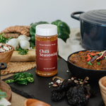 A bottle of Clean Monday Meals Chili Seasoning stands on a dark slate board surrounded by ingredients and cooked dishes, including a black pot, beans, green onions, and chili peppers, in a styled kitchen setting.