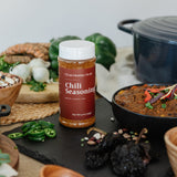 A bottle of Clean Monday Meals Chili Seasoning stands on a dark slate board surrounded by ingredients and cooked dishes, including a black pot, beans, green onions, and chili peppers, in a styled kitchen setting.