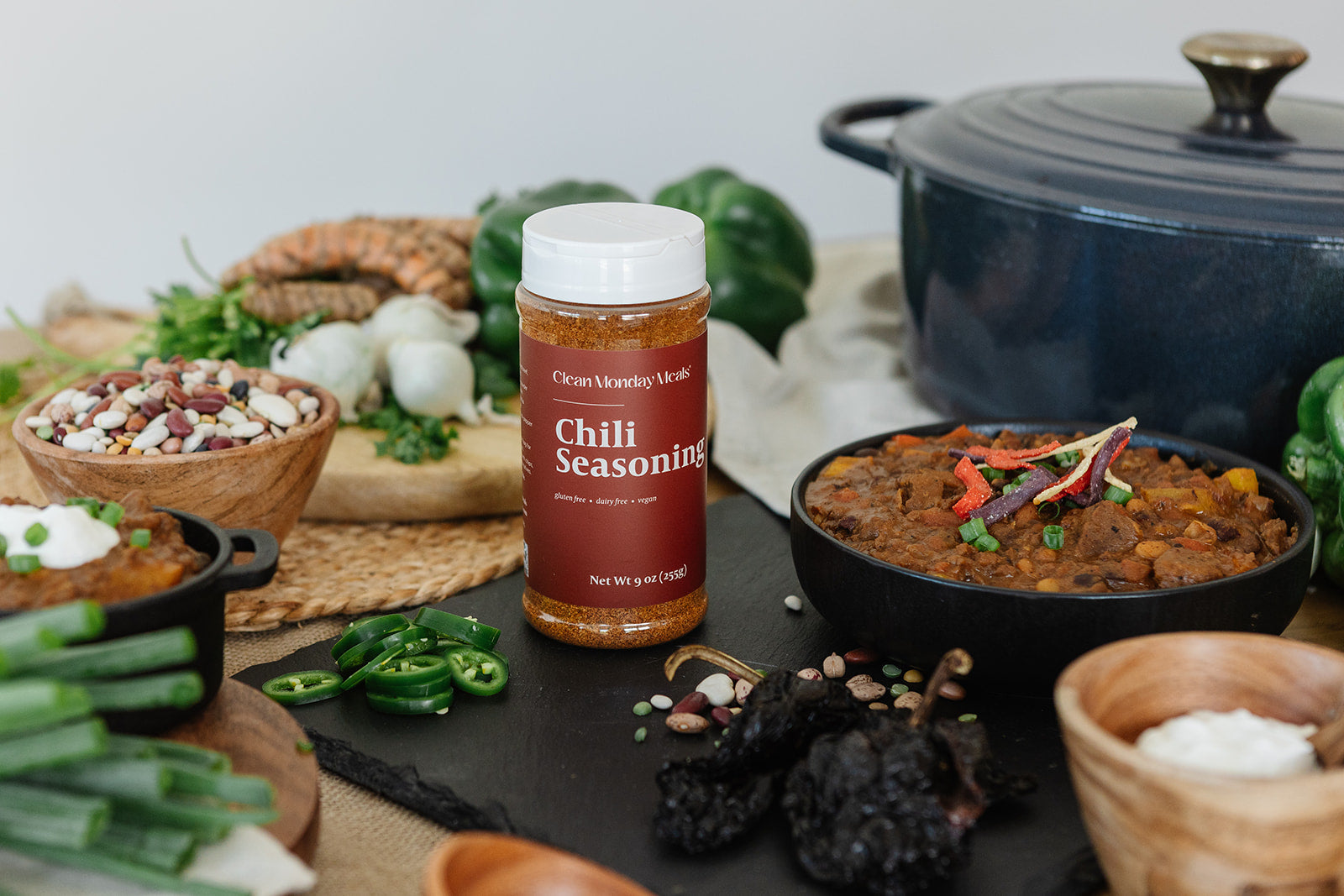 A bottle of Clean Monday Meals Chili Seasoning stands on a dark slate board surrounded by ingredients and cooked dishes, including a black pot, beans, green onions, and chili peppers, in a styled kitchen setting.