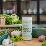 A bottle of Cream of Celery Soup Mix by Clean Monday Meals stands on a wooden cutting board surrounded by fresh ingredients like celery, garlic, red onions, and green beans in a kitchen setting.
