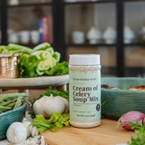 A bottle of Cream of Celery Soup Mix by Clean Monday Meals stands on a wooden cutting board surrounded by fresh ingredients like celery, garlic, red onions, and green beans in a kitchen setting.