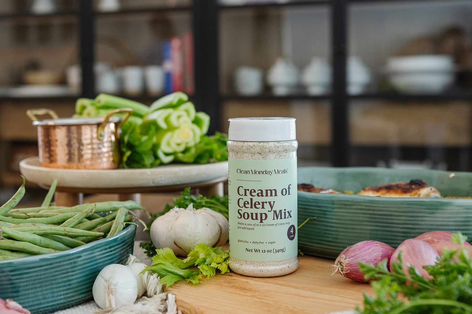 A bottle of Cream of Celery Soup Mix by Clean Monday Meals stands on a wooden cutting board surrounded by fresh ingredients like celery, garlic, red onions, and green beans in a kitchen setting.