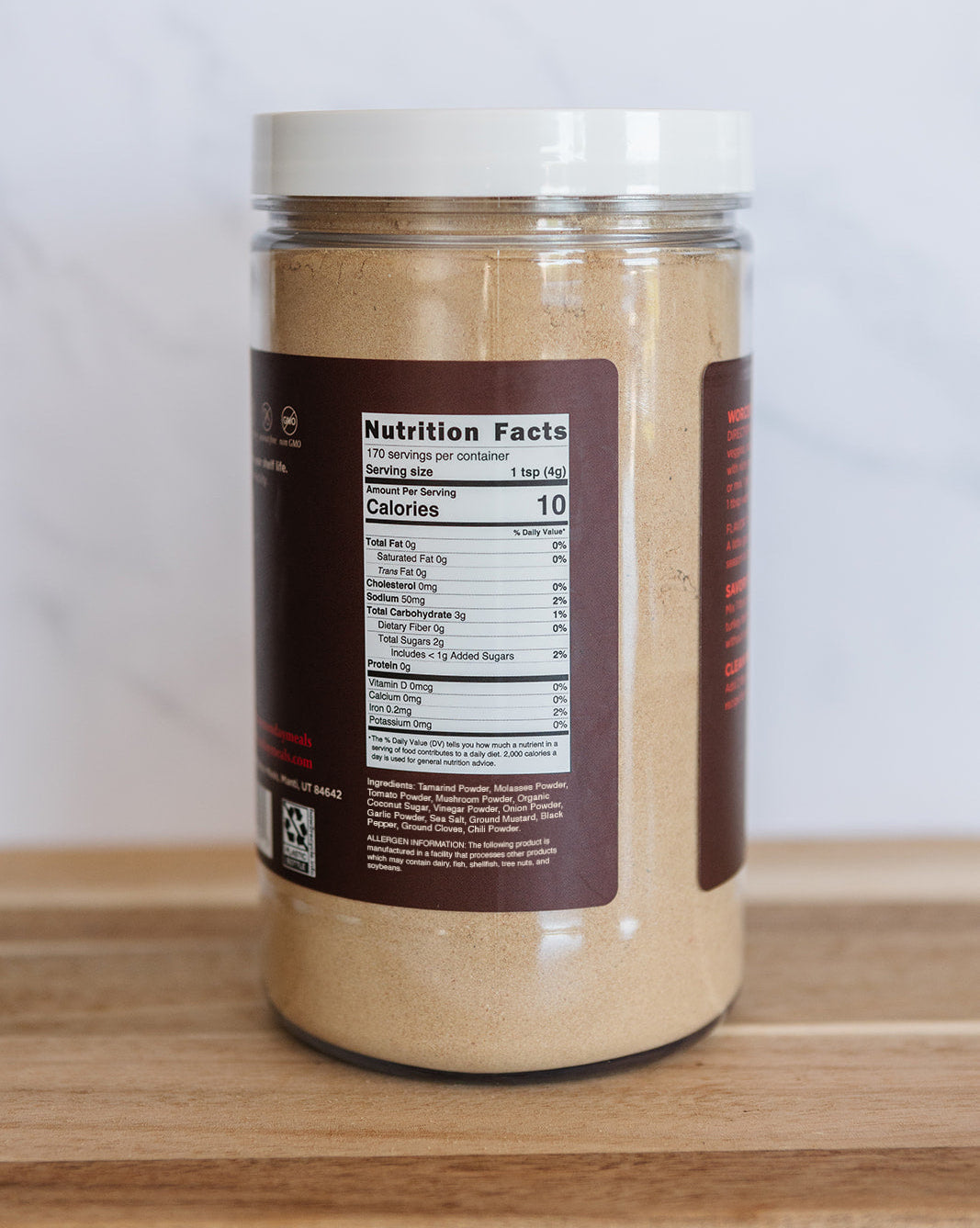 A clear plastic jar filled with a light tan powder, featuring a white lid and a dark brown label showing nutrition facts, set on a wooden surface against a light gray background.
