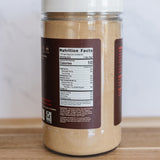 A clear plastic jar filled with a light tan powder, featuring a white lid and a dark brown label showing nutrition facts, set on a wooden surface against a light gray background.