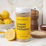 Bottle of Lemon Pepper Seasoning with lemons and a wooden pepper grinder on a kitchen counter.