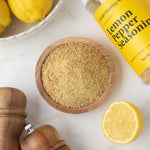 Wooden bowl of lemon pepper seasoning with a container labeled 'Clean Monday' on a marble surface with lemons.
