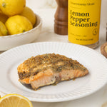 Salmon fillet on a plate with lemon pepper seasoning, lemons, and a bowl of breadcrumbs.