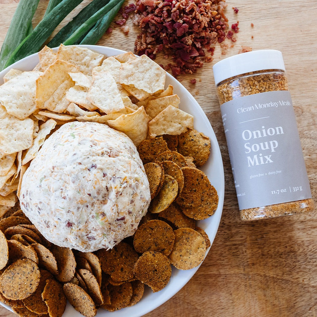 A picture of the Clean Monday Meals bottle of Onion Soup Mix next to a white plate with a Cheese ball directly in the center surrounded by chips for dipping. Above the plate sprinkled on for decoration is some bacon bits and onions.